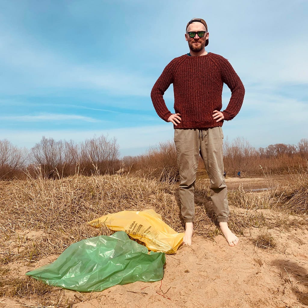 Prezes Fundacji Leśnodzieło, w czerwonym swetrze, stojący boso na plaży obok kolorowych worków ze śmieciami, symbolizujących akcję sprzątania środowiska i dbania o czystość natury.